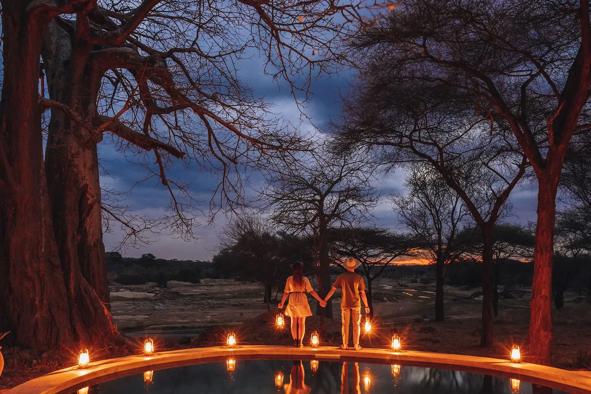 Couple holding hands by a lantern-lit plunge pool at dusk overlooking a river valley, Tanzania