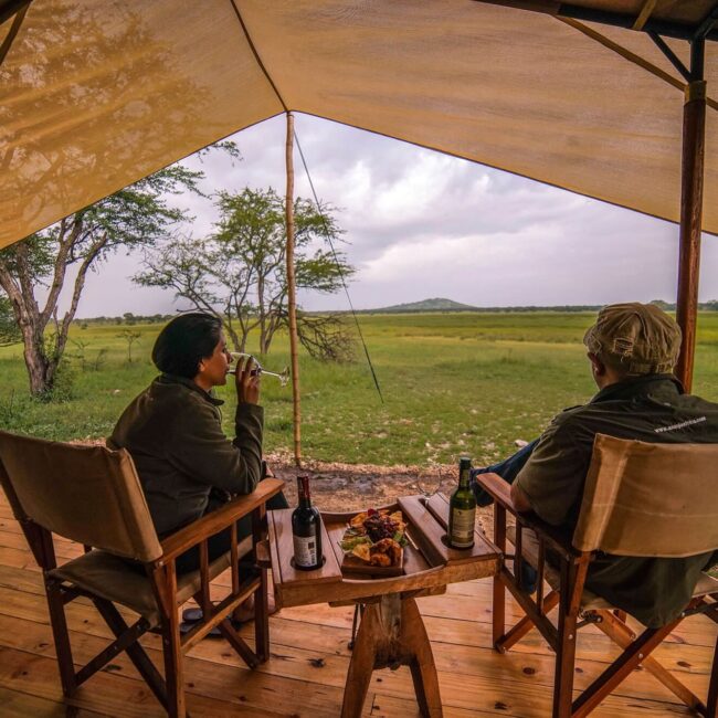 Couple enjoying wine and snacks on a tent deck overlooking the green Serengeti plains, Tanzania