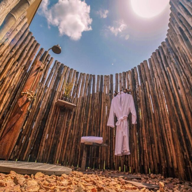 Open-air bush shower enclosed by a wooden fence under blue sky at a Serengeti safari camp, Tanzania