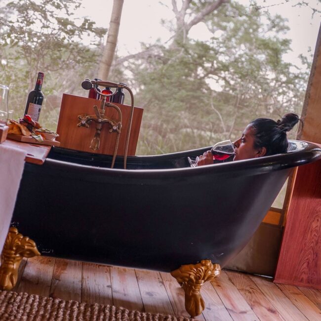 Woman relaxing with wine in a freestanding clawfoot bathtub overlooking the bush, Serengeti, Tanzania
